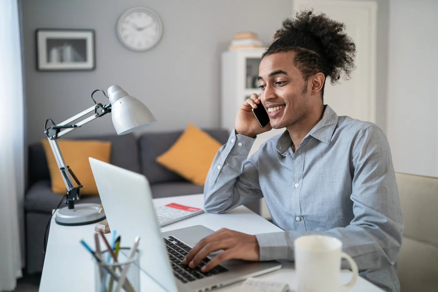 Young man holding mobile phone and talking, while typing on laptop and working at home