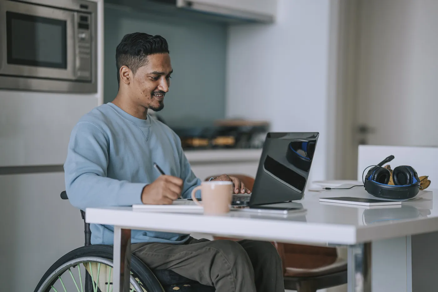 Man in wheelchair sitting at dining room with laptop and writing notes