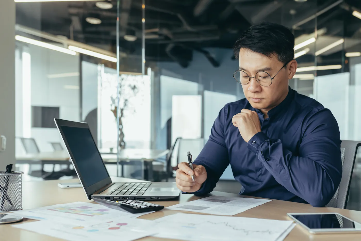 Thinking man in glasses doing paperwork, sitting at desk inside office building.