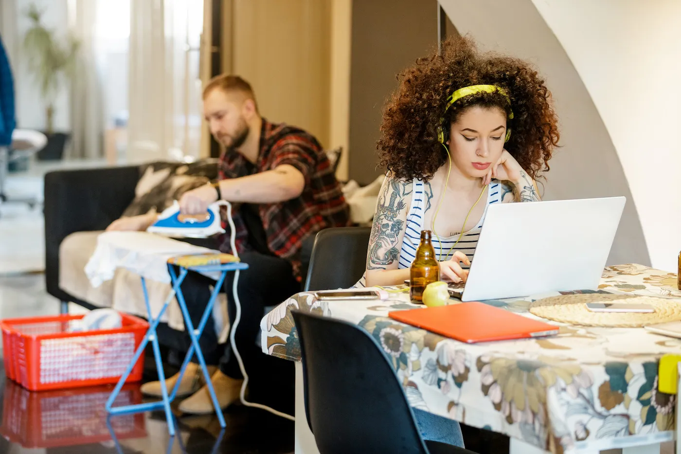 Young tattooed woman with headphones working on laptop at home, her flatmate ironing in the back