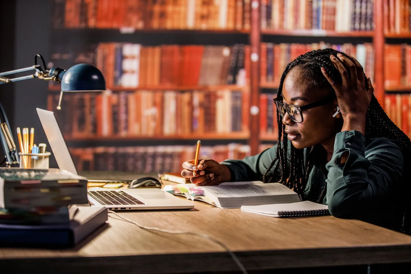 Hardworking young woman sitting at table in a library, head in hand, listening to music via blue-tooth headphones and studying for exam.