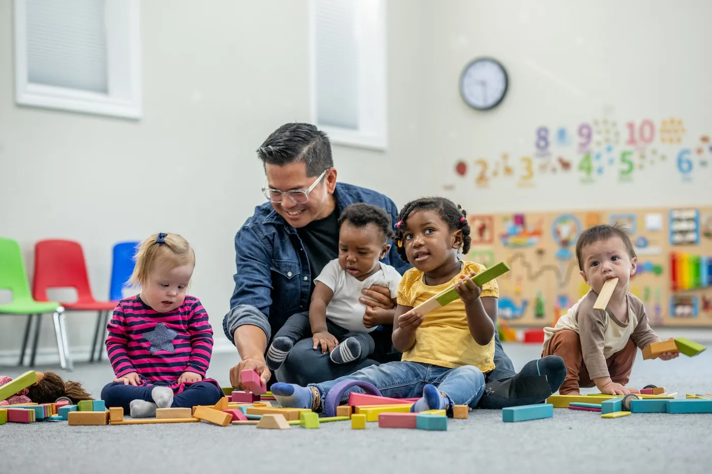 A small group of daycare children sit with their male teacher as they play together with colorful building blocks.