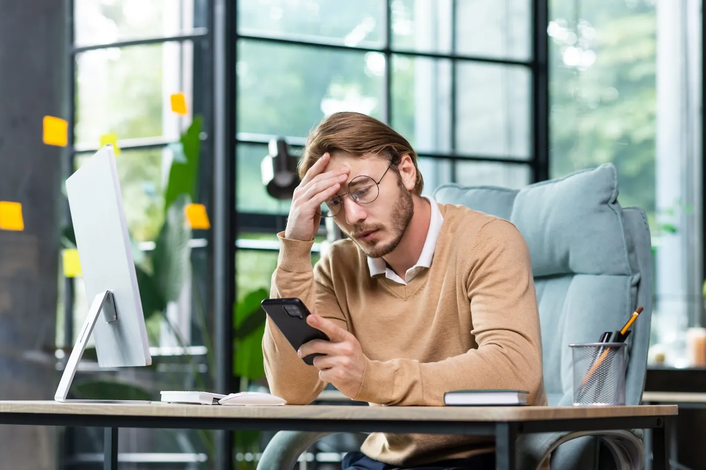 A man sits at a desk and looks at the phone. Worriedly holding his head.