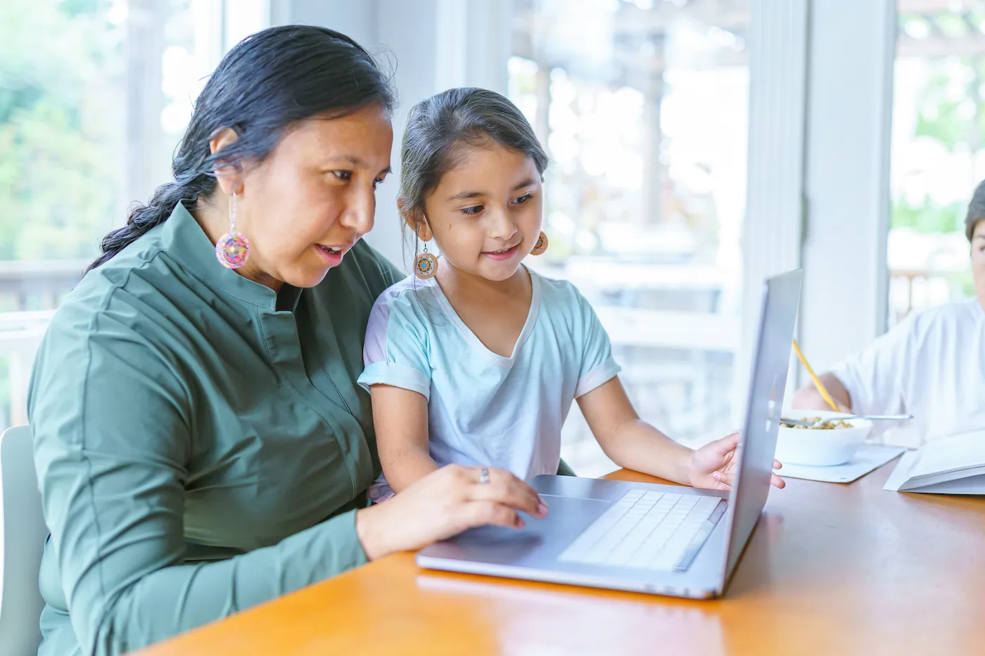 A Native American woman sits at the dining room table and works on a laptop computer as her adolescent son works on homework next to her. The woman's elementary age daughter is sitting on her lap and is looking at the computer screen.