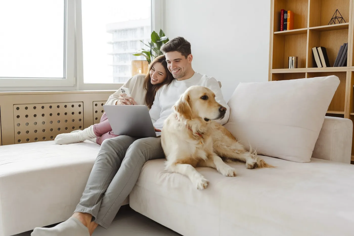 Portrait of happy young couple using laptop, hugging dog, sitting together on comfortable sofa at home.