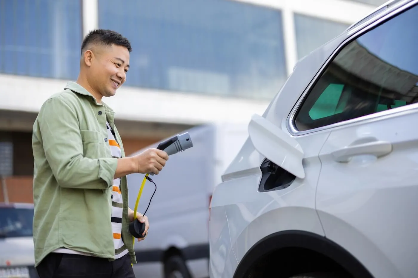 Smiling man charging an electric car using a plug-in charger outside a commercial building