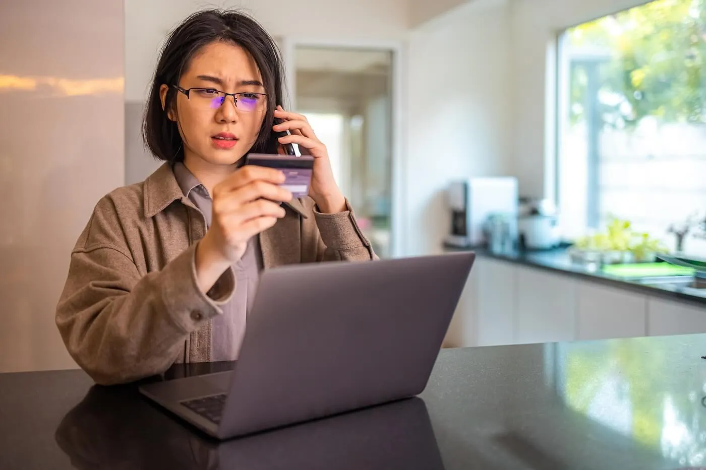 Concerned woman is making a phone call while holding her credit card, with a laptop on the desk