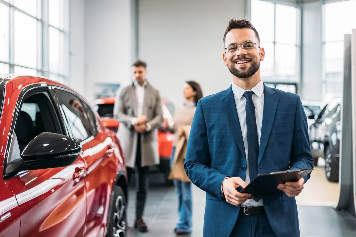 Smiling dealership agent holding a notepad, a couple is looking at the red car on the background