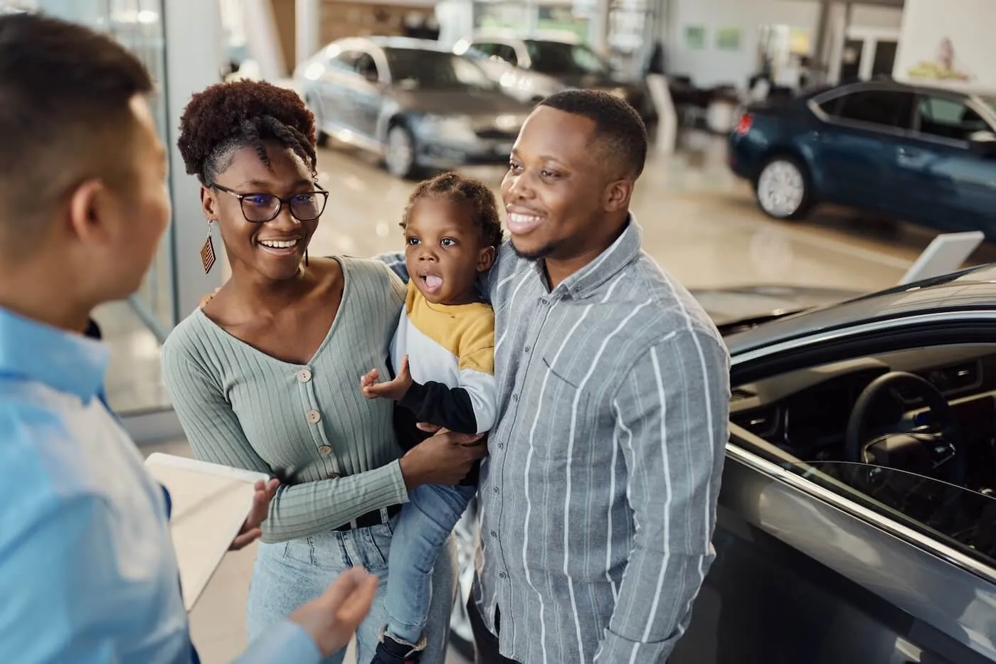 A happy family with a toddler girl are chatting with an agent in a car dealership