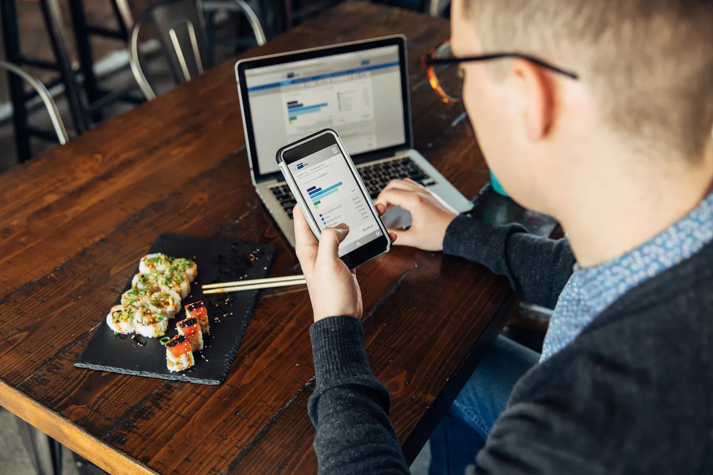 A man looking at his financial statement while eating lunch at a local sushi restaurant. He is using his bank's app on his smart phone and laptop.