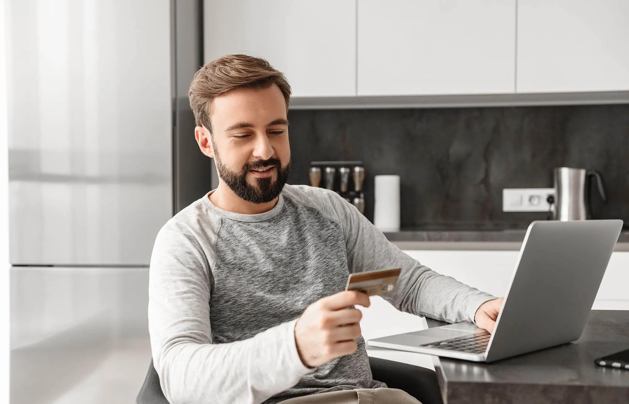 A smiling man is looking at his credit card while working on the laptop