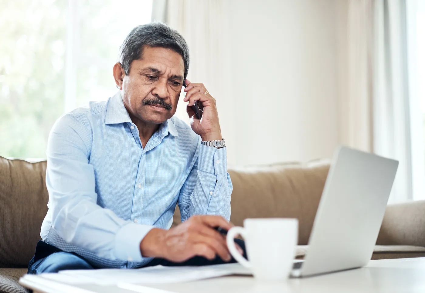 Shot of a senior man sitting on the couch, on the phone while looking at his laptop