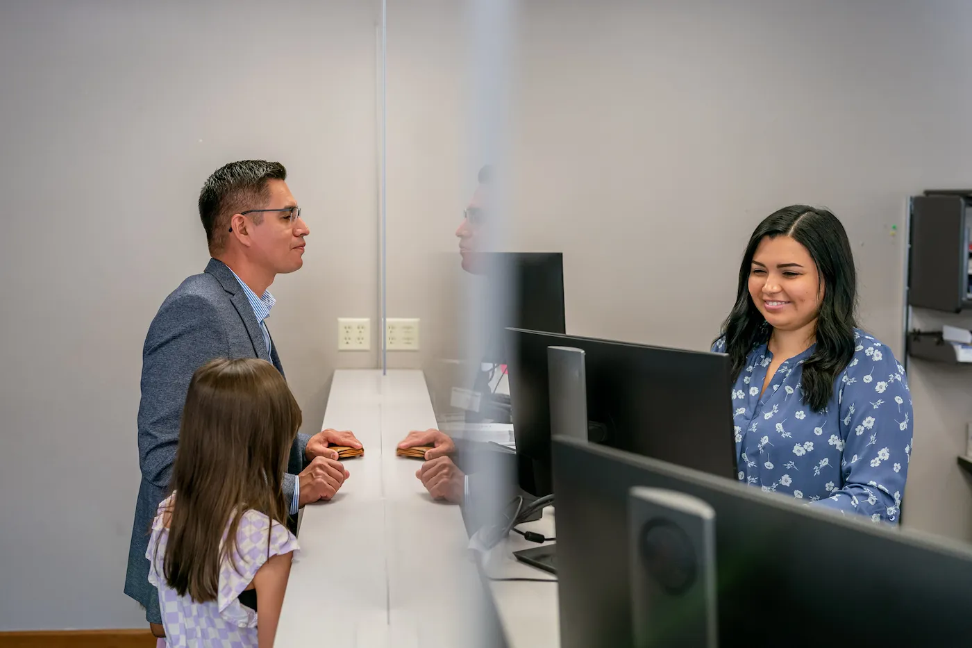 A customer service representative assists a man and his elementary age daughter at the reception desk of a bank.