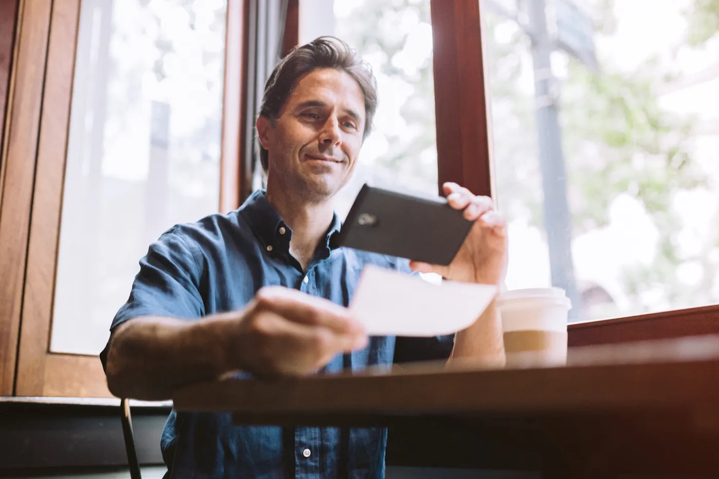 A mature smiling man in his mid 40's takes a picture with his smart phone of a check or paycheck for digital electronic depositing, also known as "Remote Deposit Capture". He sits with a cup of coffee at a cafe.