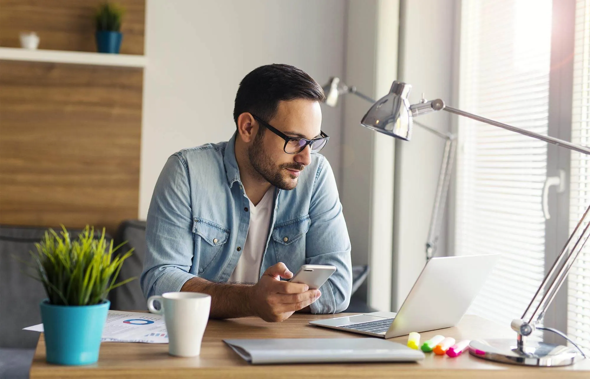 A man wearing glasses and a blue shirt looks at his laptop computer while sitting at his office.