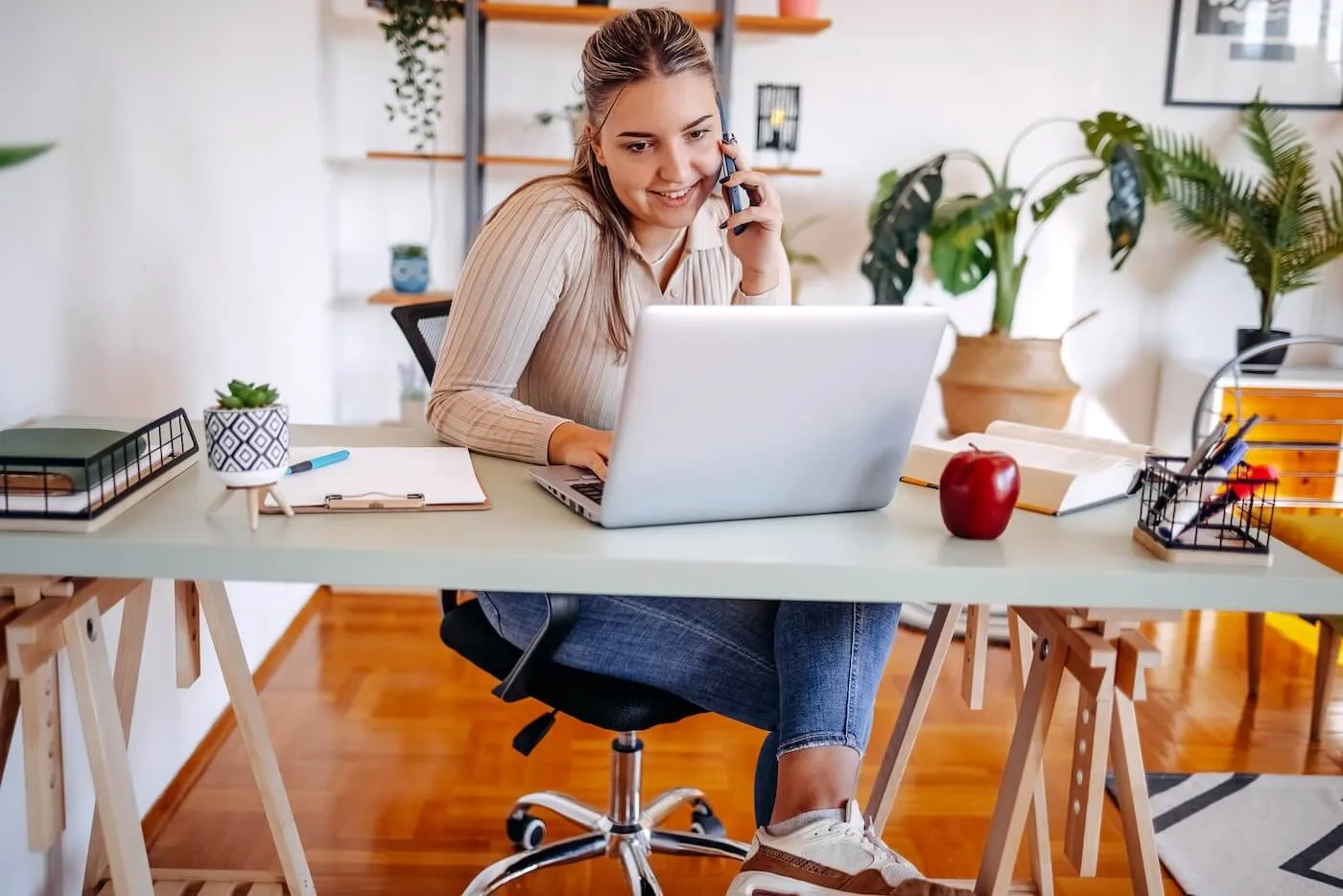 Smiling young woman is making a phone call while using her laptop in a home office