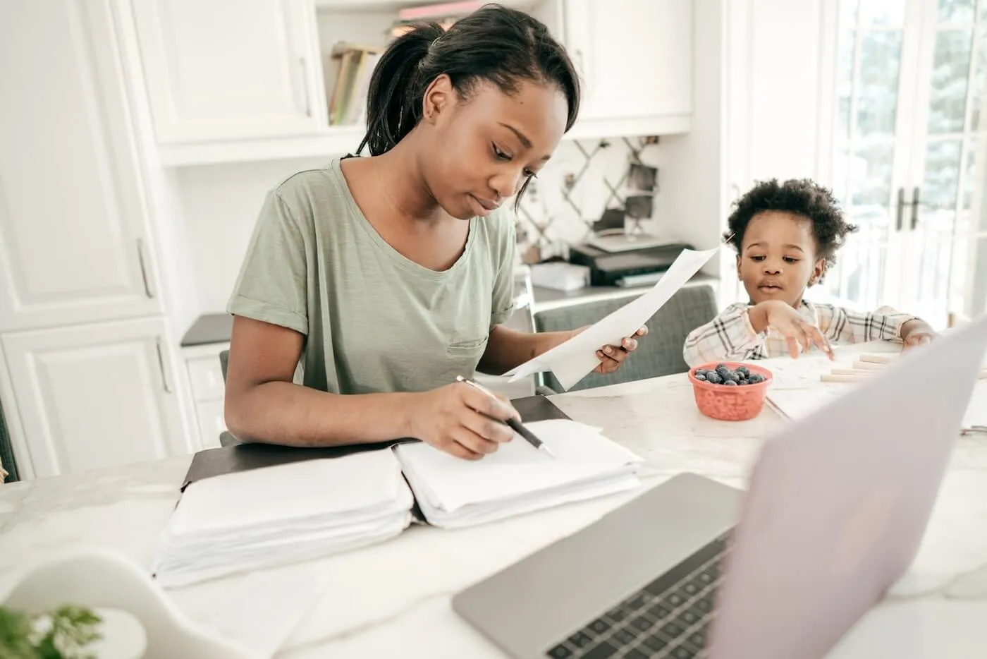 A woman is calculating taxes in the kitchen while her young kid is eating blueberries