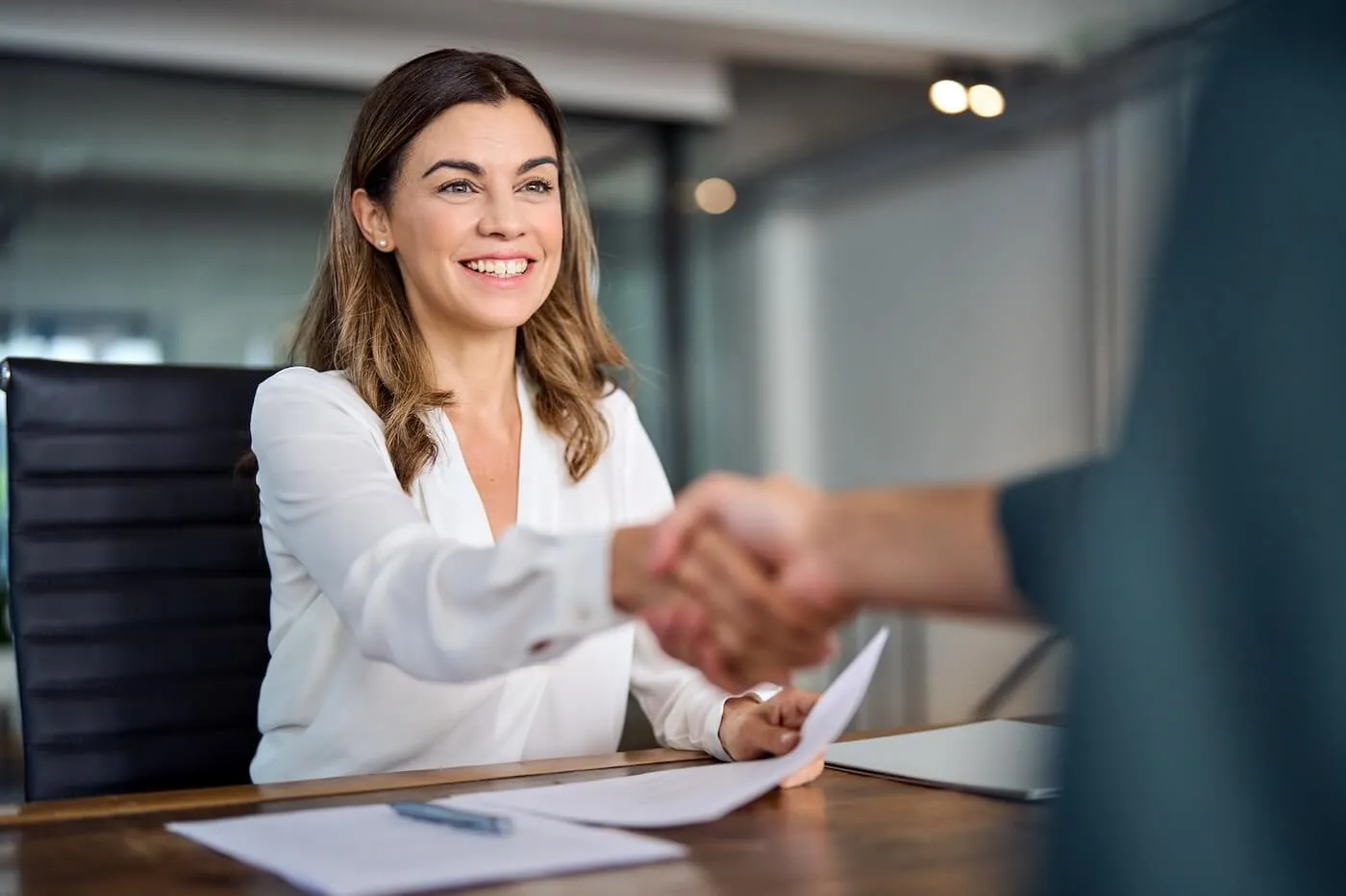 Smiling woman in business outfit holding a document and shaking hands with another person in the office