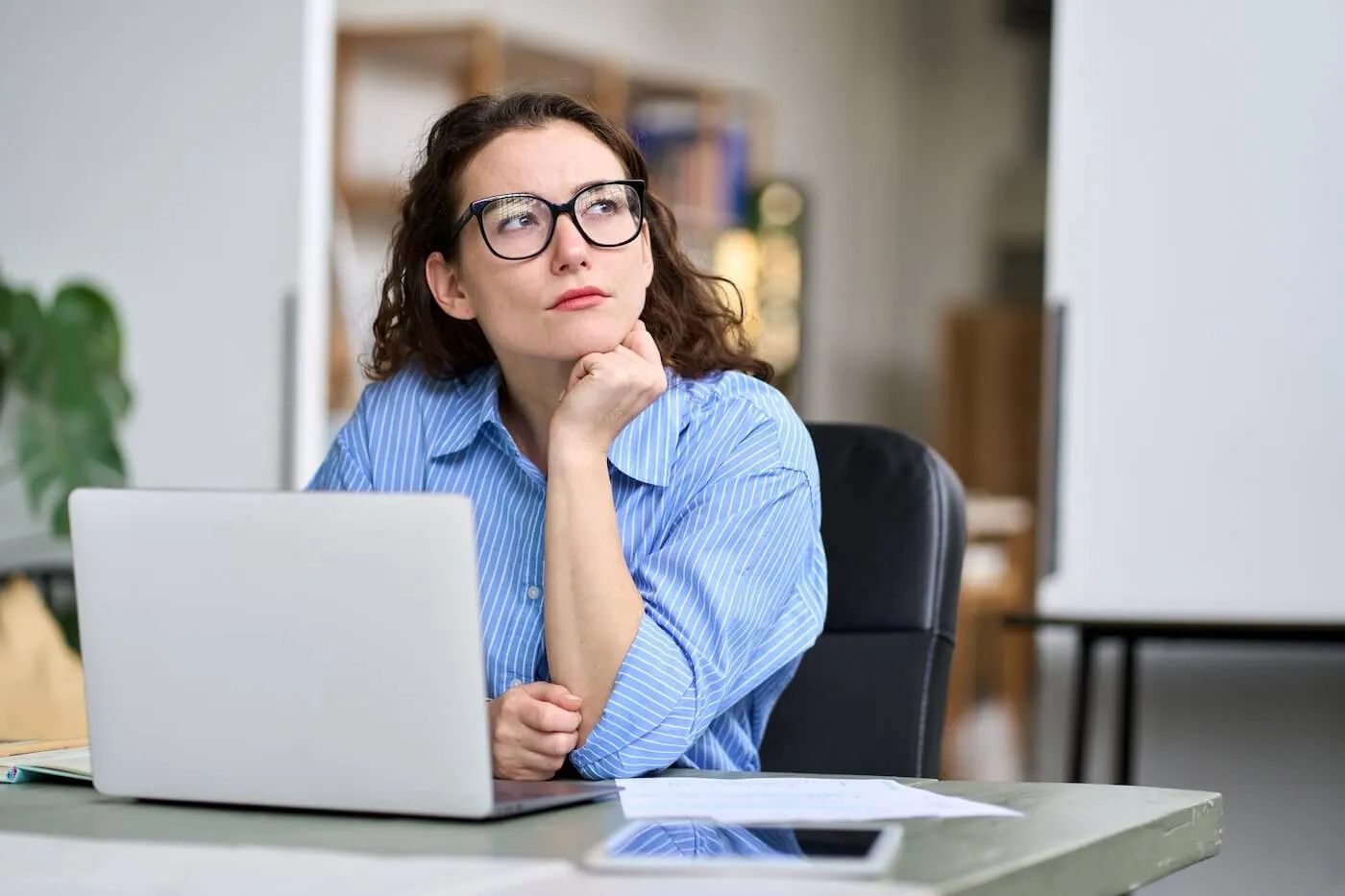 A thoughtful woman looks into the distance with an open laptop on her desk