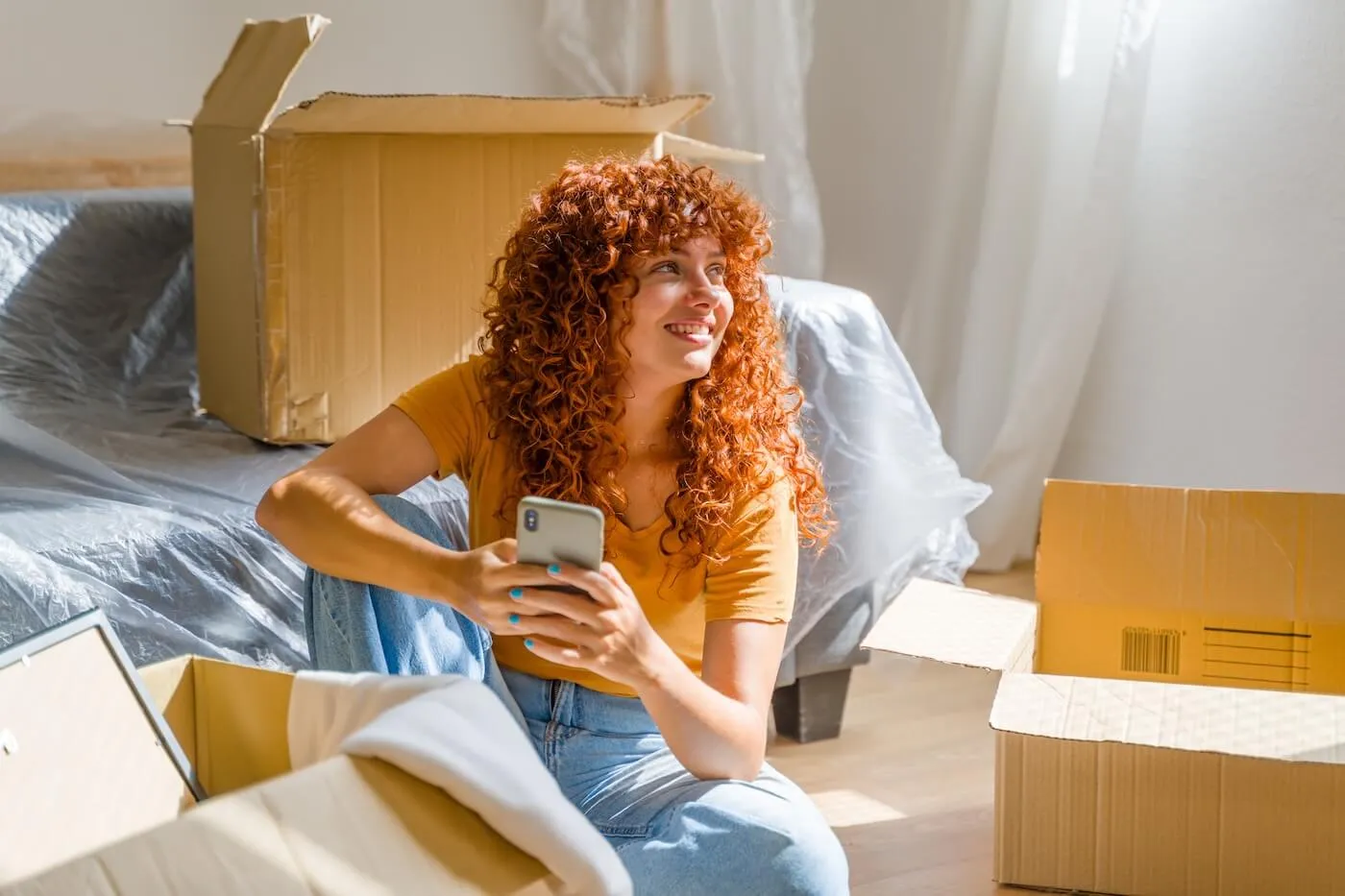 Smiling woman with curly ginger hair is sitting on the flor of her new living room and using her smartphone, surrounded by moving boxes