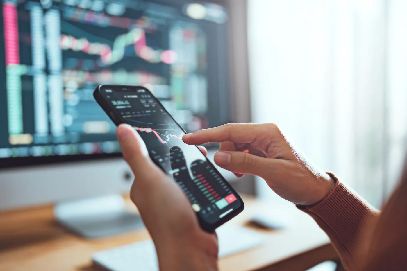 A woman checking her investments on her phone with another investment chart open on her desktop computer.