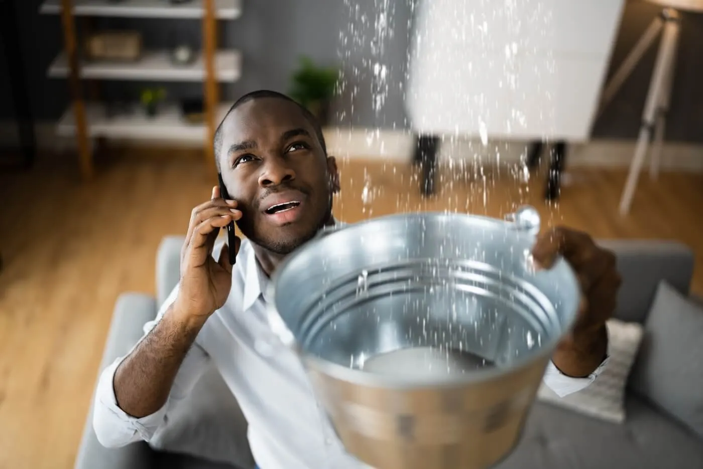 A concerned man is making a phone call while holding a bucket under a ceiling leak