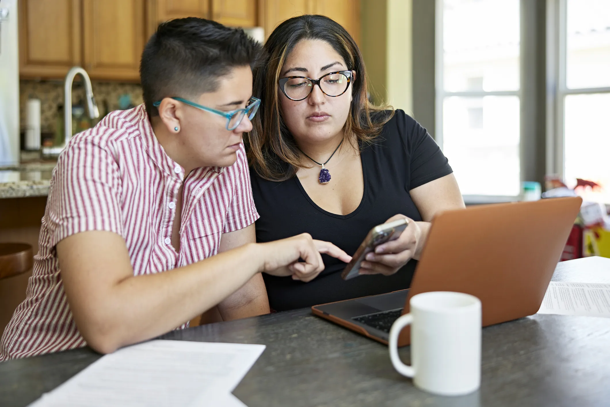 Waist-up view of women in early 30s sitting together in family home with savings account paperwork, laptop, and making calculations on smart phone.