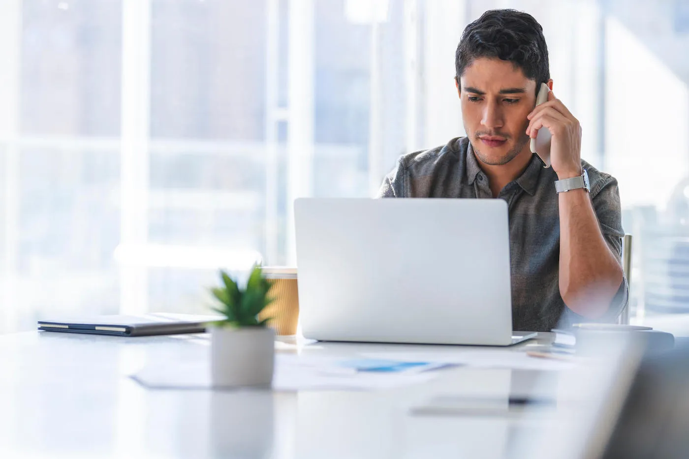 Serious young man holding phone looking at laptop screen.