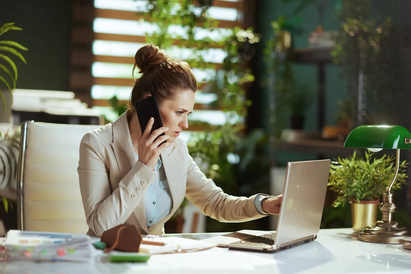 Woman in a light business suit in modern green office with laptop speaking on a smartphone.