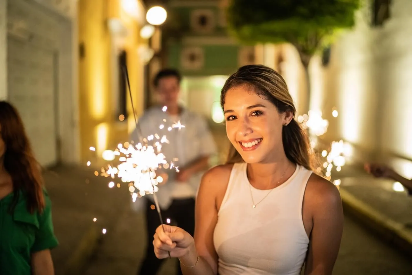 Smiling young woman in a tank top holding a sparkler while walking on night street. More people are celebrating on the background.