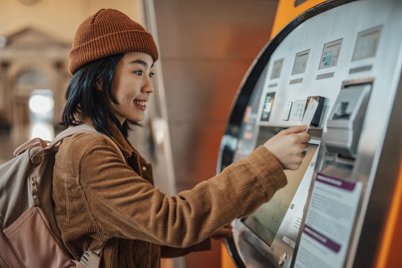 A traveler wearing a backpack withdraws money from an ATM in a train station.