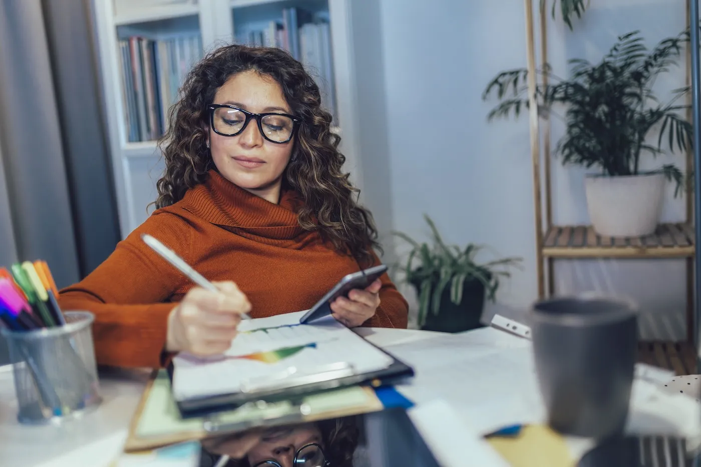 Woman holding phone in her hands while taking notes, calculating family finances at home.