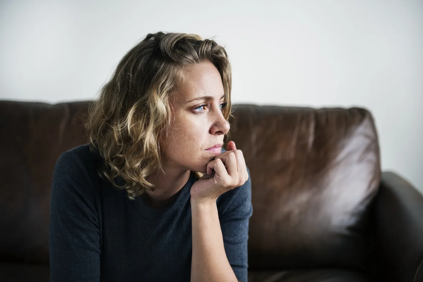 A pensive woman looks out a window while sitting on a leather couch.