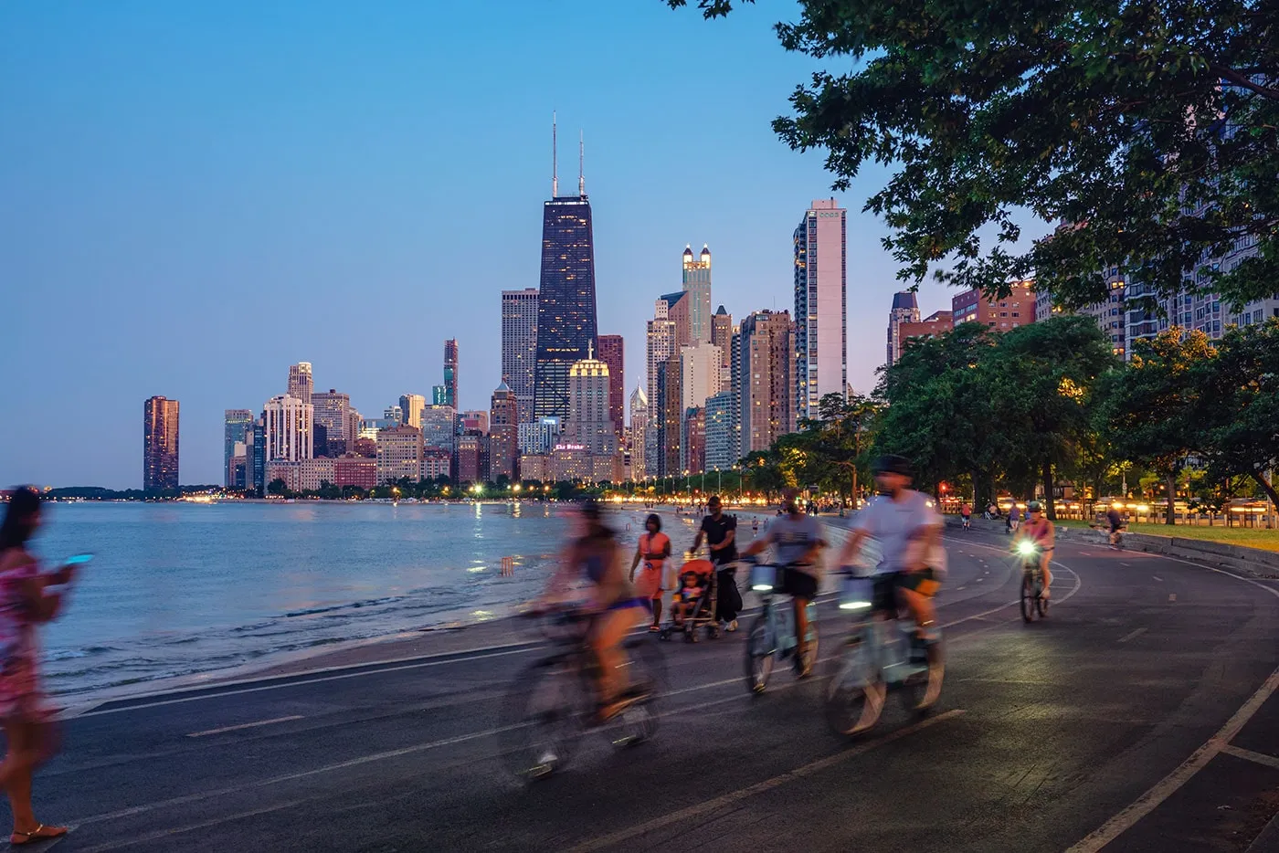 Pedestrians and bicyclists are on the road next to a lake with the city skyscrapers in the background.