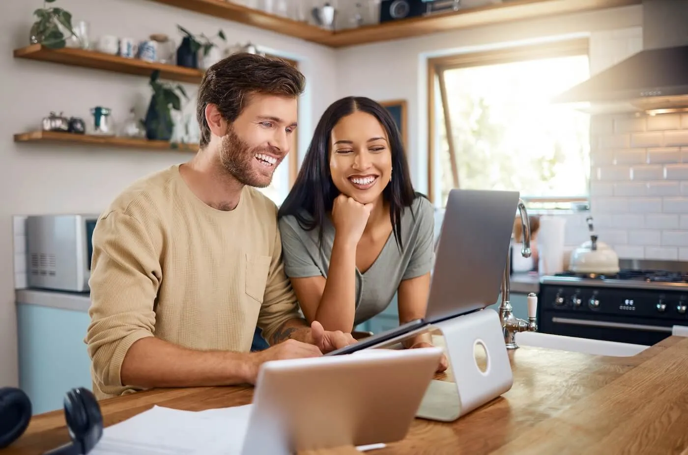Happy couple standing at a kitchen counter and looking at a laptop placed on a stand, with another open laptop in the foreground; wooden shelves with dishes, a stove, and a kettle are visible in the brightly lit kitchen background.