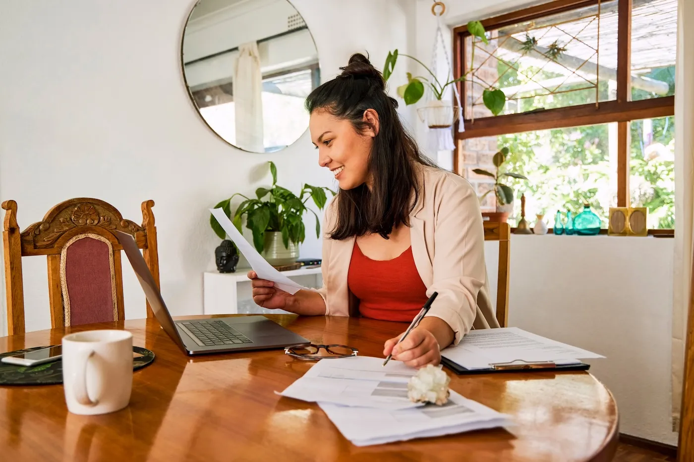 Shot of a young woman using her laptop and checking paperwork at home