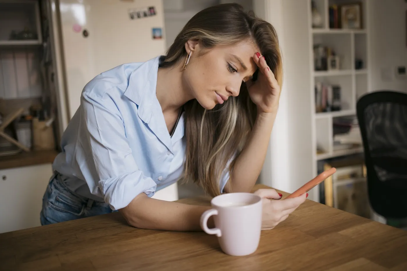 Young woman using her cellphone while drinking coffee in kitchen.