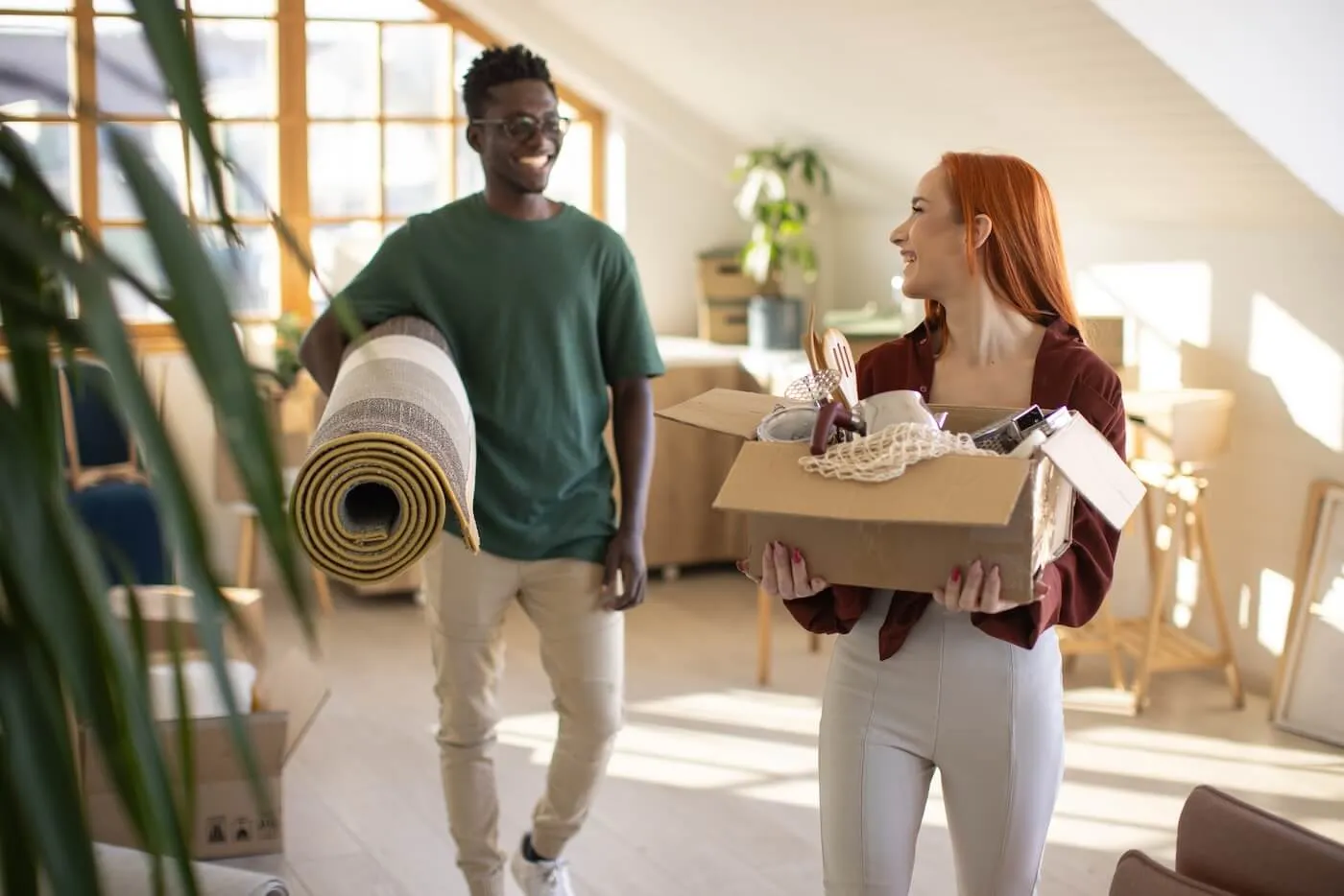 Happy young couple moving into a new home, with the woman carrying a moving box and the man carrying a rolled‑up carpet