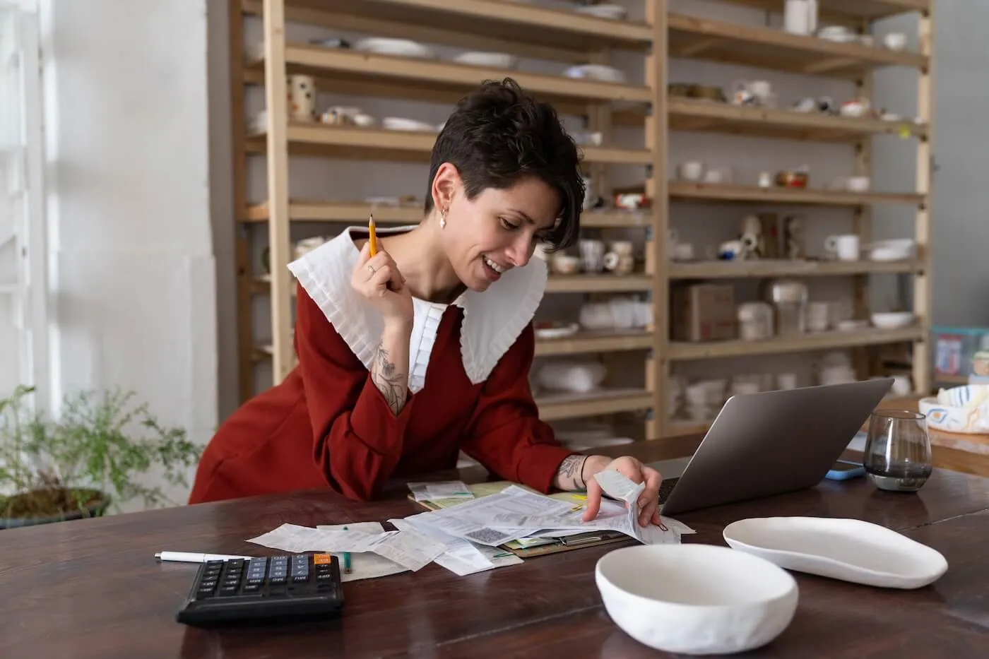A young woman reviewing receipts and notes at a desk with a laptop and calculator, focused on organizing finances, with shelves of ceramic goods in the background.