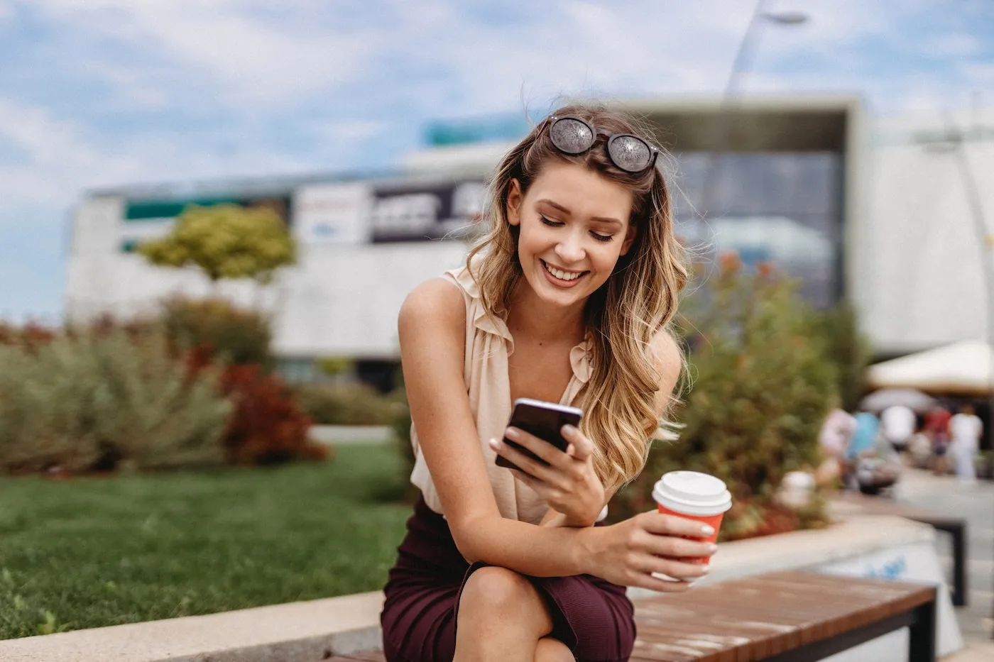 A woman sitting outside on a bench, holding her coffee and smiling at her phone.