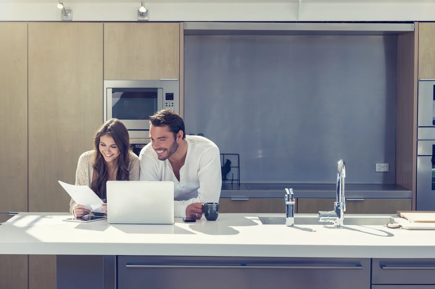 Happy couple with paperwork. They are standing at the kitchen bench working online with a computer and bills. They have coffee.