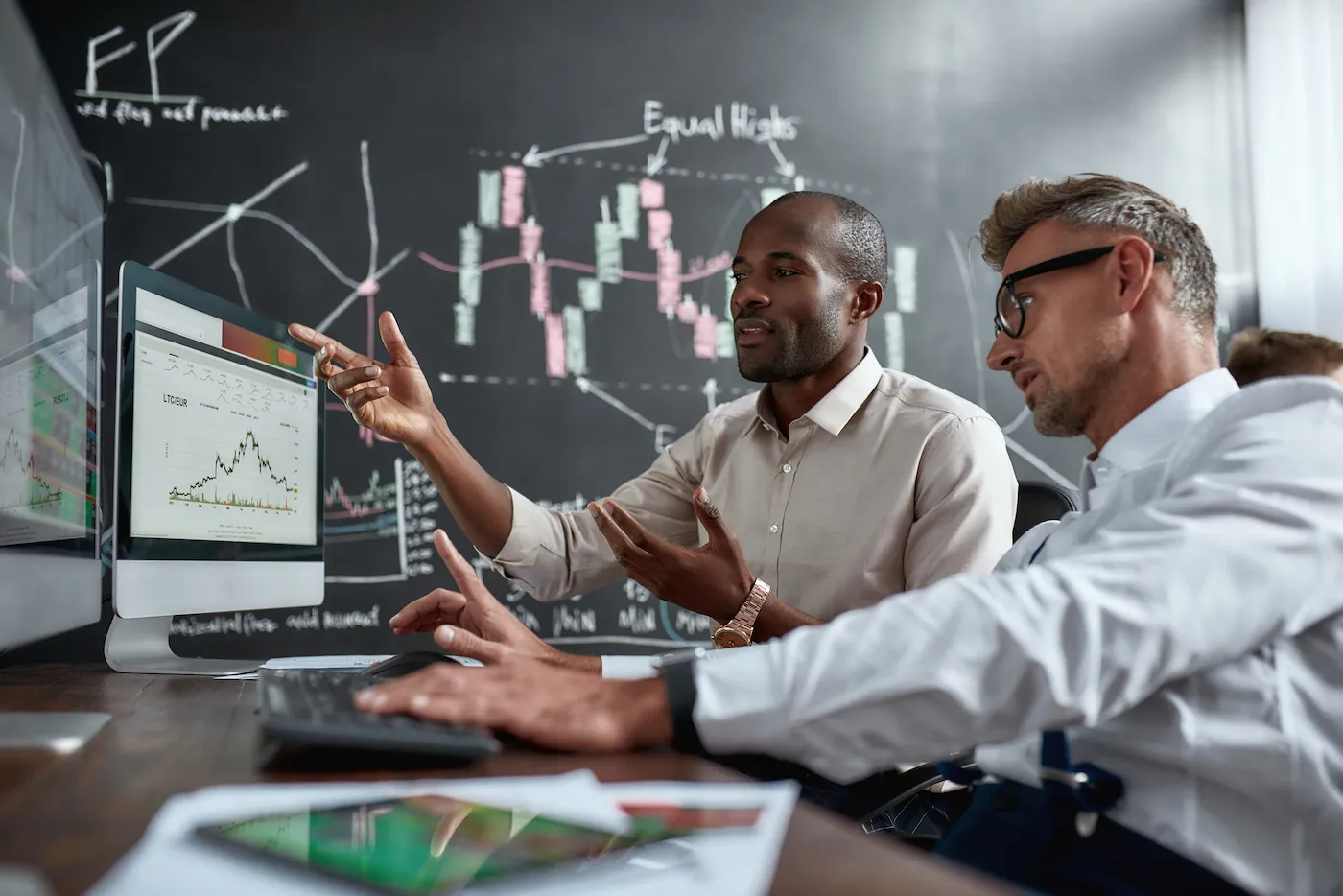 Two colleagues traders talking to each other, looking at graphs while sitting in the office in front of multiple computer screens.