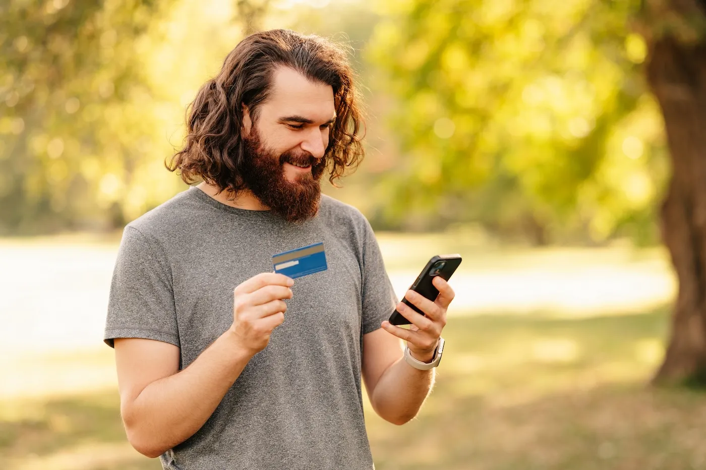 A bearded man is using his phone and credit card while buying online, standing in a park.