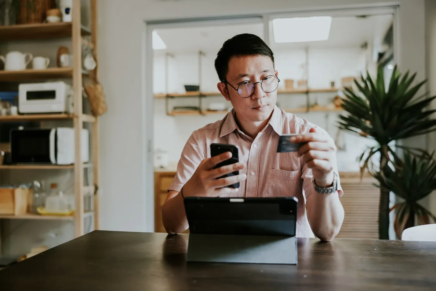 A man using his credit card and phone in front of his laptop. He is sitting at the dining table in his home.