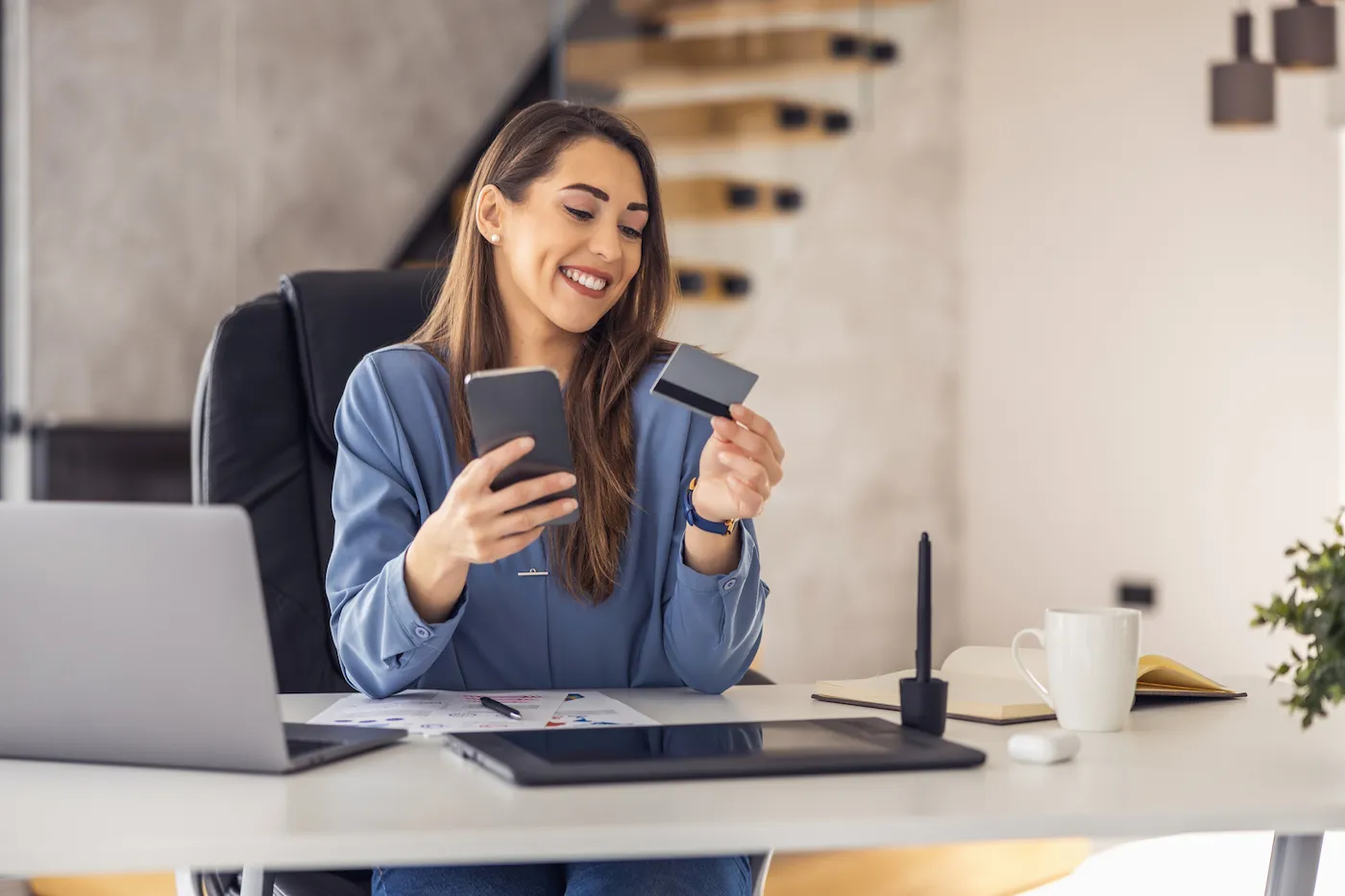 Shot of young woman working with laptop in the office, holding her credit card in one hand and her phone in the other.