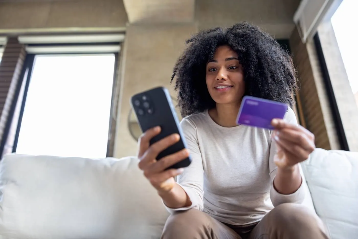 Smiling young woman seated on a couch indoors holds a smartphone in one hand and a payment card in the other, with a window, and a neutral-toned interior visible in the background.