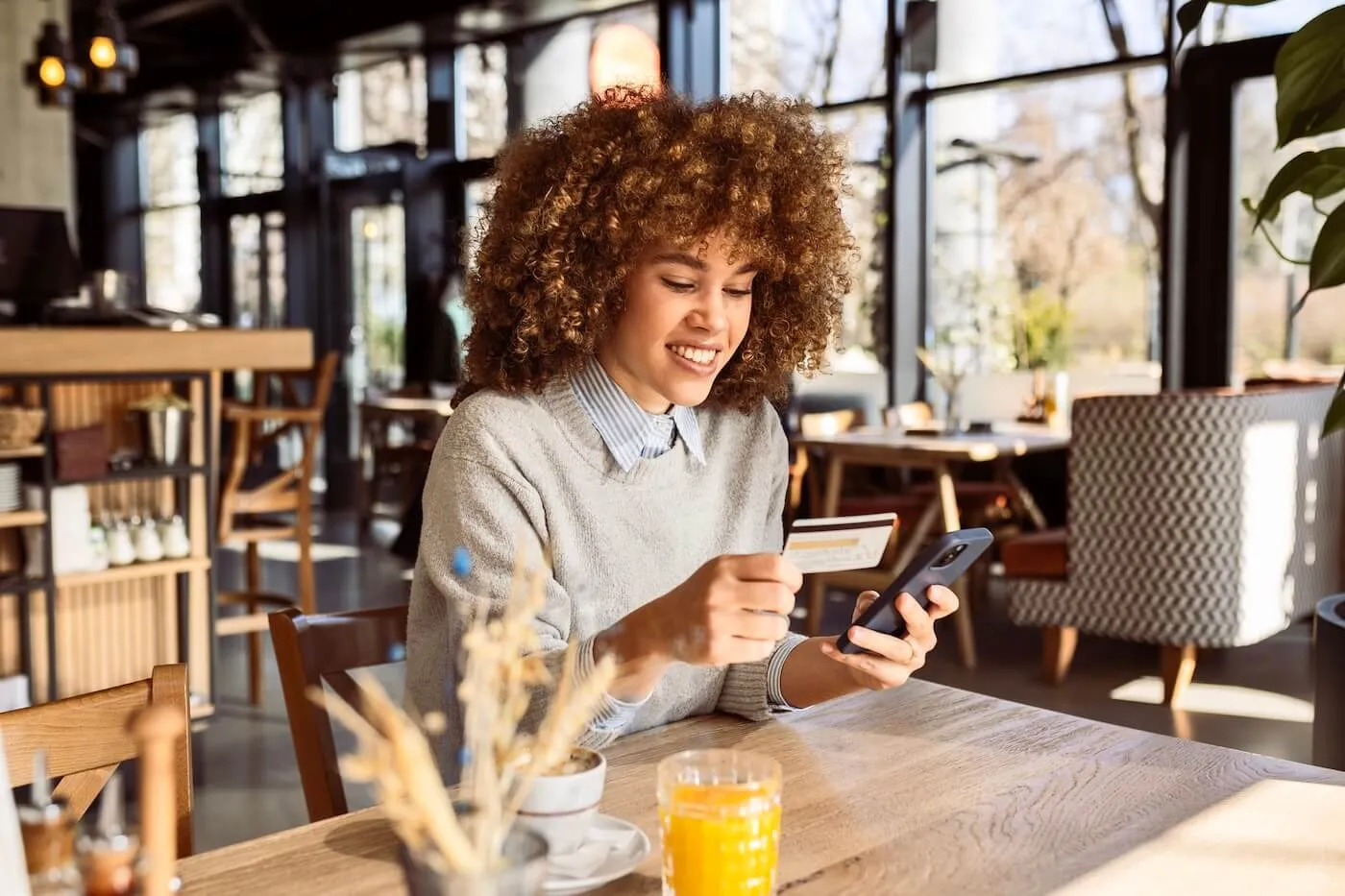 Smiling young woman seated at a café table uses a smartphone while holding a payment card, with a glass of orange juice, a cup and saucer, and small tabletop items visible, and modern café furniture, large windows, and indoor plants in the background.