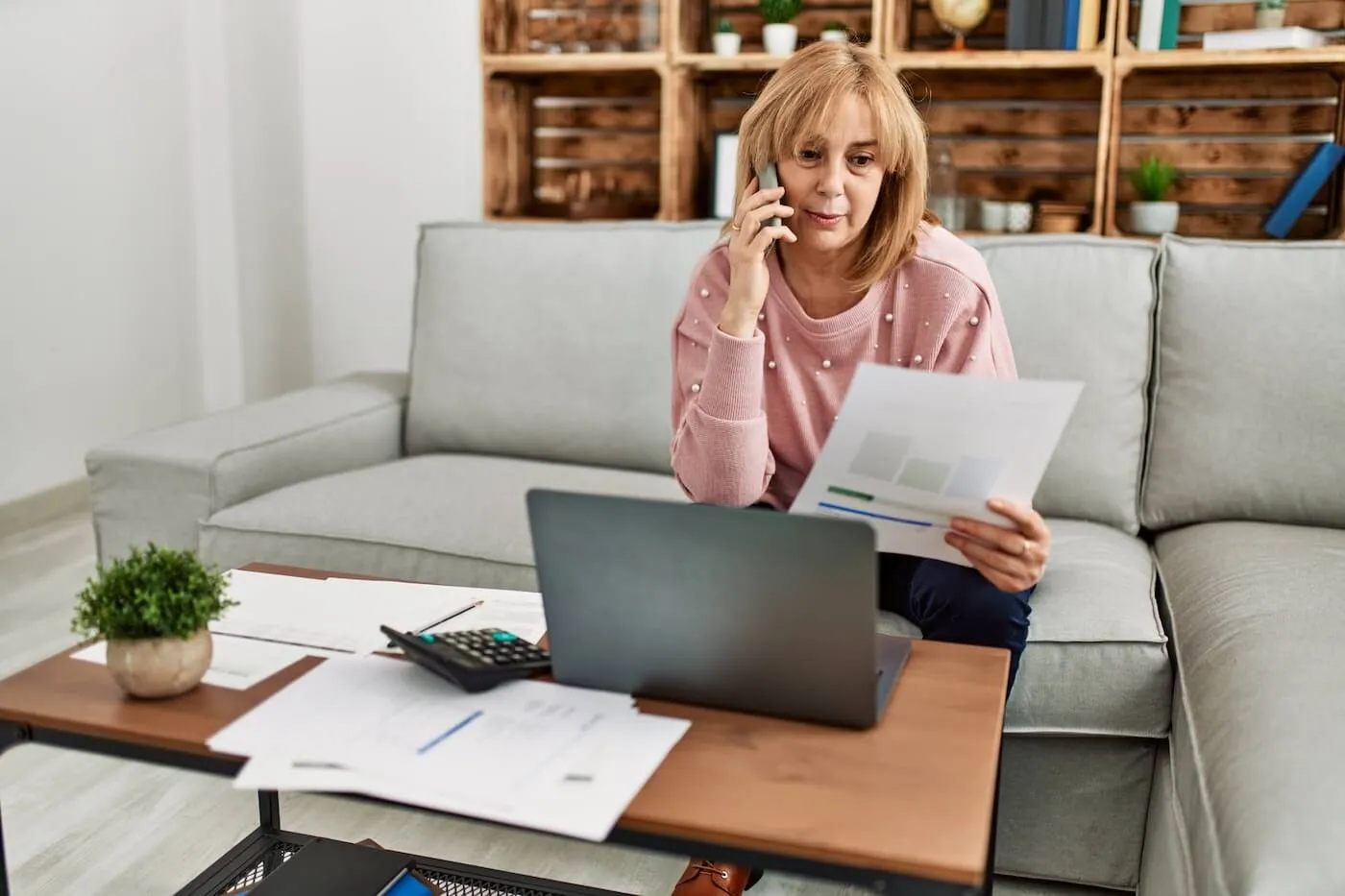 A mature woman is calling the lender to discuss the debt settlement while looking at her bills. She sits on the sofa next to the coffee table with more printouts, a calculator, and an open laptop.