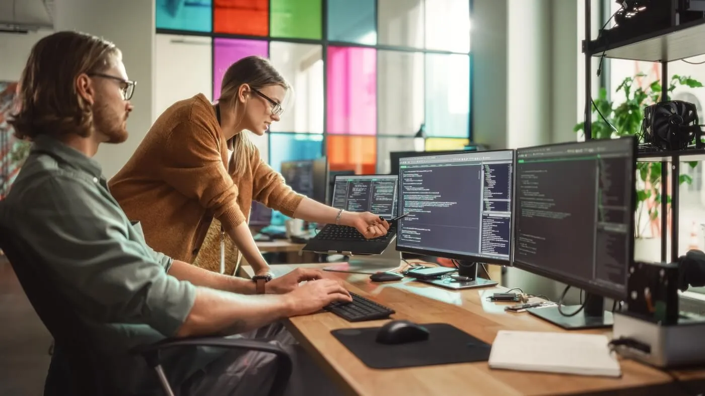 A couple of coders, male and female, reviewing the code on the monitors in the sunny office with a stained glass window