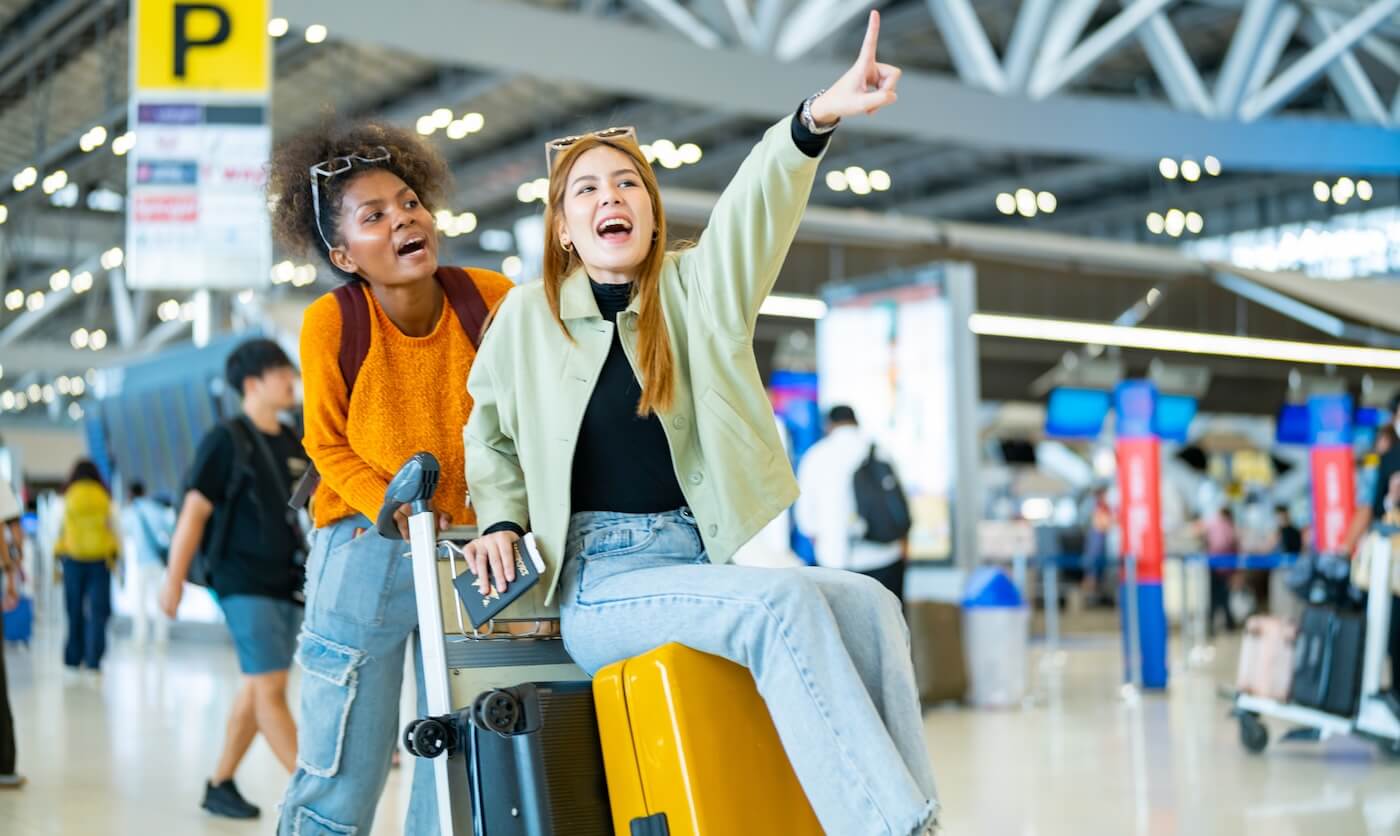 Two happy female friends in an airport terminal; one is sitting on a suitcase and pointing at something with excitement
